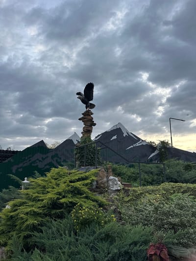 The image depicts an outdoor scene, likely a park or decorative garden area, with a sculpture of an eagle perched atop a stack of stones. In the background, there are stylized mountain-like structures made of painted wood or metal, suggesting a themed environment. Lush green vegetation, including bushes and trees, fills the foreground and middle ground. The sky is overcast with dramatic, cloudy formations, hinting at either pre-storm conditions or twilight. The overall impression is of a man-made landscape designed for aesthetic appeal, possibly within a public space. The location is identified as Yaroslavskaya, Russia.