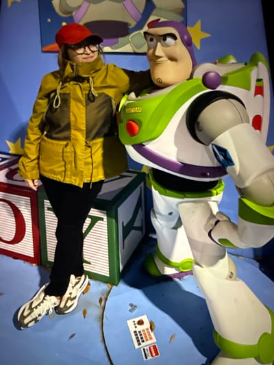 An individual wearing a red cap, glasses, and a yellow jacket stands with their left hand on the shoulder of a Buzz Lightyear costume. The Buzz Lightyear costume is positioned to the right of the individual and appears to be a prop or display. Both subjects are standing in front of a blue backdrop with painted yellow stars and a large character image at the top. In the foreground, there are large, colorful blocks that resemble toy building blocks, one with a large red "O" and another with a green border and a white "A". The ground is a blue surface with scattered brown leaf-like debris. Two small white cards with text are visible on the blue ground to the right of the individual's feet. This scene appears to be a photo opportunity or display, likely within an indoor entertainment venue in Taipei, Taiwan, possibly related to a theme park or promotional event.