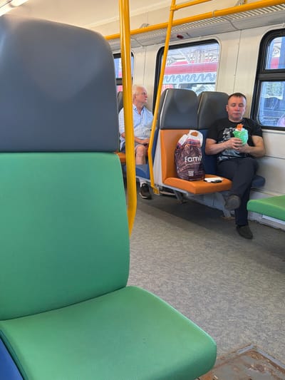 The image shows the interior of a train carriage. In the foreground, a green seat is visible. In the middle ground, a man wearing a black t-shirt and dark pants is sitting on an orange and blue seat, holding a green object wrapped in plastic. To his left, a shopping bag with the word "Fami" is placed on the seat. Further in the background, an elderly man with white hair, wearing a light blue shirt and shorts, is seated. The train carriage appears to be modern with yellow poles and windows showing a view of another train and station platforms, suggesting it is located in Podolsk, Russia. The floor of the carriage is a speckled grey material.