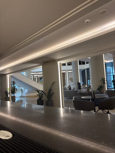 The image shows a modern hotel lobby with a minimalist design. A polished gray counter runs along the bottom foreground, reflecting the ambient lighting. Behind the counter, a sleek, white staircase ascends to an upper level. The lobby features tall, fluted pillars and recessed lighting, creating a sophisticated atmosphere. Several potted plants are strategically placed, adding a touch of greenery. In the background, a seating area with comfortable chairs and tables is visible, with a few patrons casually interacting. The overall scene suggests a contemporary and upscale environment, consistent with a hotel lobby in a modern city like Kazanlak, Bulgaria.