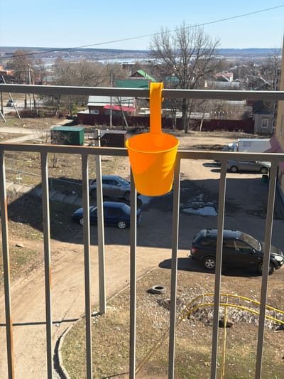 The image captures an outdoor scene, likely viewed from an elevated position such as a balcony, looking down and across a residential area. In the immediate foreground, a bright yellow plastic funnel or scoop is attached to a gray metal railing, which shows signs of rust in places.
Beyond the railing, the view extends over an unpaved, dirt ground that serves as a parking area. Several cars are visible: a dark-colored sedan is parked on the right, and further left, a silver hatchback and a blue sedan are also parked. The ground is dry, with patches of sparse grass and scattered debris, including a discarded tire and a yellow flexible pipe or hose near the dark sedan. The presence of strong, elongated shadows suggests a sunny day, likely mid-morning or afternoon, under a clear blue sky.
In the mid-distance, numerous trees with bare branches indicate that the season is late winter or early spring. Scattered among these trees are various residential buildings with different colored roofs, including red and green. Further in the background, a wide, lighter-colored expanse is visible, possibly a river or a large body of water, which could be partially frozen or reflecting the sky. Distant hills or land formations frame the horizon. A faint wisp of smoke or haze can be seen above the distant trees. The overall setting depicts a somewhat semi-urban or rural residential landscape, consistent with Sarapul, Russia. There are no people visible, and no discernible text. The scene is static, offering a serene observation of the surroundings.