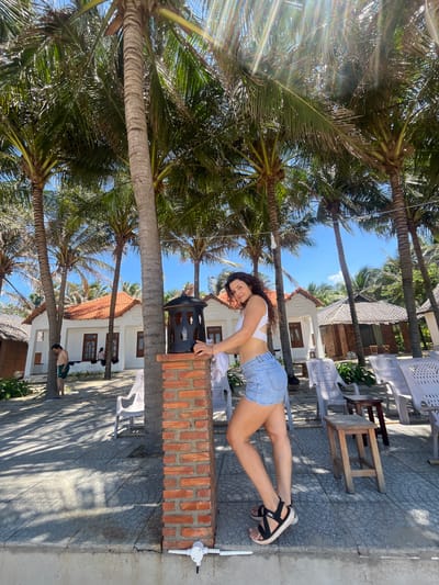 The image captures a sunny day in Mui Ne, Vietnam, likely at a resort or beachfront area, indicated by the presence of palm trees and small bungalow-style buildings with orange tiled roofs. A young woman with curly brown hair, wearing a white bikini top and denim shorts, stands in the foreground, leaning against a brick pillar adorned with a black lantern. She is looking towards the camera with a slight smile. Behind her, several white plastic chairs and small wooden stools are arranged on a paved area. In the background, one of the bungalow buildings is visible, with a man in green shorts standing near a palm tree. The scene is bathed in sunlight, casting shadows from the palm trees onto the ground.
