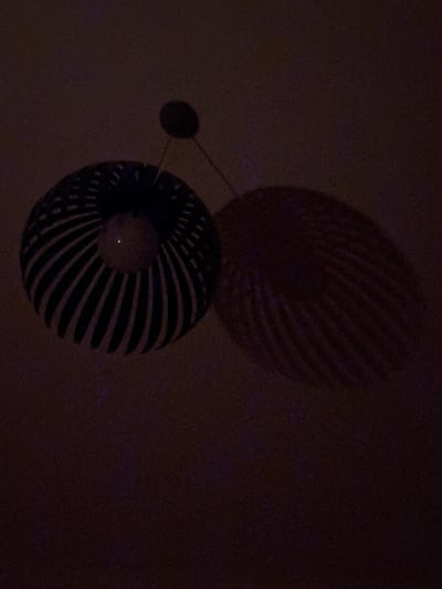 An underexposed, medium shot captures a decorative lamp hanging from a ceiling. The lamp shade is a dark color with spiraling white stripes. A bright, small light emanates from the center of the lamp. A shadow cast by the lamp is visible on the wall behind it, mimicking the lamp's shape. The environment is dark, suggesting nighttime or a room with low lighting. The image is too dark and lacks identifying features to determine its specific location within Nesebar, Bulgaria.