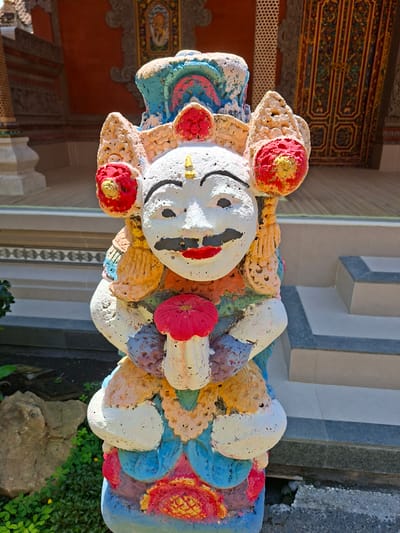 The image shows a colorful statue of a jovial figure, likely a religious or cultural icon, situated in an outdoor setting. The statue has a round, white face with black eyes, a small gold nose, and a red mouth. It is adorned with various textures and colors, including shades of blue, red, yellow, and orange. The statue appears to be made of a porous material, possibly stone or concrete.
The backdrop reveals architectural details, including a wall with vertically alternating gray and white striped columns, and ornate patterns on the ceiling. There is foliage around the statue and stone or concrete structures in the background.
Given the artistic style and architectural elements, it's reasonable to suggest the statue is located in Ubud, Indonesia, which is known for its rich cultural heritage and artistic expressions.
