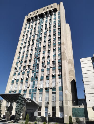 The image depicts the Moldtelecom headquarters building in Chișinău, Moldova. It is a tall, Brutalist-style concrete building with a facade of numerous windows and air conditioning units. The sky is clear blue, and a few birds are visible flying in the distance. In the foreground, there is an entrance area with a modern canopy, some trees, and signage that reads "Moldtelecom". To the right of the main building, another building with a similar architectural style is partially visible. The scene is set in an urban environment, bathed in bright sunlight.