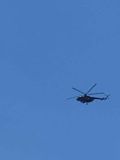 A dark-colored helicopter with its rotors spinning is captured mid-flight against a clear blue sky. The helicopter is positioned in the right half of the frame, angled slightly downwards. The image does not provide any identifiable landmarks or context to suggest a specific location within the city of Novosibirsk, Russia, or any other city. The scene is devoid of people, interactions, or any environmental features other than the sky.