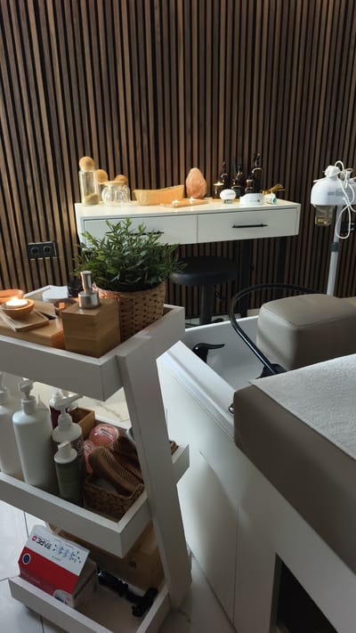 This image depicts a spa treatment room in Kuşadası, Turkey. The scene is set against a backdrop of dark wooden slatted walls. On a white vanity table, several items are arranged, including decorative glass bottles, a large pink salt lamp, a woven basket, and small lit candles, suggesting a relaxing ambiance. A black stool is positioned in front of the vanity.

In the foreground, a white multi-tiered trolley holds various spa products. The top shelf contains a potted green plant in a wicker basket, a wooden tray with a lit candle, and a wooden dispenser. Below this, more bottles and a woven basket with combs are visible. The bottom shelf displays a box labeled "BARE + FACE MASK" in red and white lettering, along with a black tool.

To the right, a white spa pedicure station is prominent, featuring a cream-colored cushioned seat and footrest, and a black faucet. A white steam machine with a flexible hose is positioned next to it. The overall setting suggests a professional spa environment designed for relaxation and beauty treatments. The time of day is not explicitly indicated, but the lighting suggests it could be evening or a dimly lit interior.