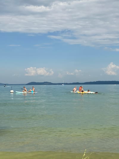 This is a wide shot of a lake in Milvignes, Switzerland, on a sunny day. In the foreground, the clear water shows a sandy bottom. In the mid-ground, two people are on a paddleboard. The person on the right is a male wearing blue swim trunks. The person on the left is a female with her hair in a bun, wearing a bikini and holding a paddle. They are facing left. Further out in the lake, another group of people are on inflatable rafts. One person is wearing an orange life jacket. In the background, a line of hills forms the horizon, and the sky is blue with scattered white clouds. There are several boats visible on the water in the distance.