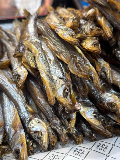 This is a close-up, overhead shot of a pile of small, dried fish arranged on a patterned surface. The fish are various shades of brown and golden yellow, with some appearing more silvery. Their bodies are elongated and slender, with visible fins and tails. The heads of the fish are pointed, and many have open mouths. The overall texture of the fish appears dry and somewhat brittle. The patterned surface beneath the fish has a repeating geometric design in black on a white background. The lighting suggests an outdoor or well-lit indoor environment. There is no indication of the city of Jerusalem, Israel in this image.