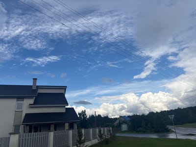 The image depicts a residential scene under a dynamic sky, likely in the Капыльскі сельскі Савет, Belarus. In the foreground, a portion of a modern house with a dark, multi-pitched roof and light-colored walls is visible on the left. A grey, picket-style fence runs across the front of the property. Behind the house, a dense line of dark green trees forms the background, obscuring the horizon in many areas. Beyond the trees, several residential structures are sparsely visible, including one with a blue roof and a smaller, light green shed-like building next to it. The sky is a prominent feature, with a bright blue expanse partially filled with scattered, wispy white clouds and larger, billowy cumulus clouds on the right. Four dark power lines stretch diagonally across the upper portion of the frame, from left to right. The road in the mid-ground appears wet, suggesting recent rainfall, with puddles visible. A triangular road sign is planted on the right side of the road. The overall atmosphere suggests a rural or suburban setting with evidence of overcast and potentially clearing weather.