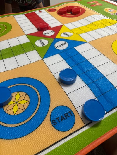 The image depicts a close-up, eye-level view of a Ludo board game. The board is colorful, with sections in orange, yellow, green, and blue, divided by white squares. Several game pieces are visible: three red circular pieces are clustered in the upper right quadrant of the board on red squares, and three blue circular pieces are present in the lower half of the board, with one on a blue circle labeled "START," and two on white squares. The lighting is bright, suggesting an indoor environment, possibly illuminated by natural light from a window or artificial lighting. The focus is sharp on the game board and pieces, with the background blurred, hinting at a person or furniture. The image does not contain any information that would place it in Marten, Bulgaria.
