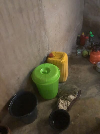 The image depicts a dimly lit corner of an interior space, possibly a storage area, with rough, textured walls and a concrete floor. In the foreground, a large black plastic basin or bucket is visible on the left, partially filled with a dark liquid. To the right of the basin, a bright green plastic bucket with a lid is situated. Behind these, a large yellow plastic jerrycan stands upright. Further to the right, a collection of bottles and containers are arranged on a shelf or surface, including a red bottle and smaller bottles with green caps. An orange-colored container and some round green objects, possibly fruits, are also present in the background. A crumpled piece of fabric or material is lying on the floor between the green bucket and the jerrycan. The overall impression is one of a functional, utilitarian space.
