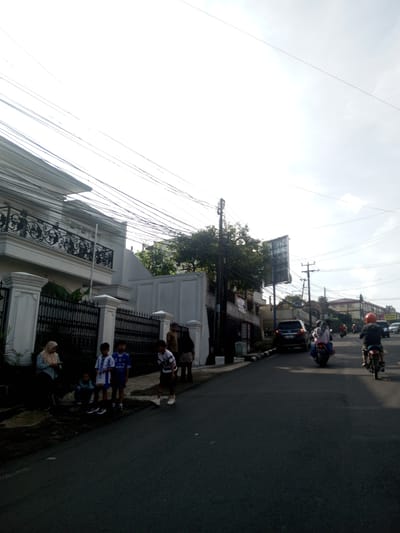 The image shows a street scene with a residential area. On the left, a group of people including children are standing by a fence in front of a white building. On the road, there is a black car and several motorcycles, suggesting traffic flow. Overhead, there are numerous power lines. The scene appears to be set during daylight under slightly overcast conditions. This image does not appear to be in Phou Khoun, Laos.