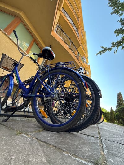 The image shows several bicycles leaning against a concrete wall. One bicycle is prominent, painted blue with a black basket attached to the handlebars. The scene appears to be outdoors, with a bright blue sky visible in the background and greenery suggesting a natural setting. A building with yellow walls and balconies is visible to the right of the bicycles. The image is taken from a low angle, looking upwards. The time of day appears to be daytime, likely sunny, given the brightness of the sky. There are no people visible in the image. The text "CITYTHINK" is visible on the blue bicycle's frame. The location is identified as Nesebar, Bulgaria. The image is oriented vertically.