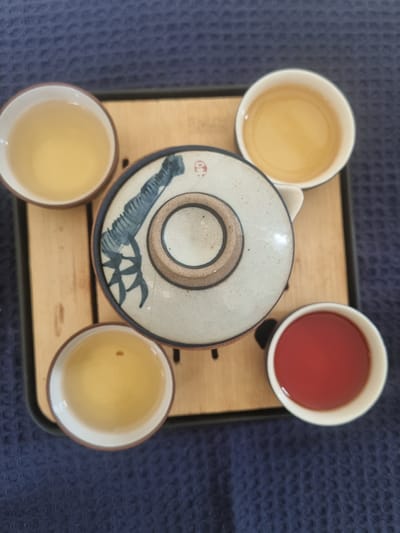 On a dark blue textured surface, a light-colored wooden tray with a black border holds a tea set. The set includes three cups filled with a light-colored beverage and one cup with a red beverage. A circular teapot lid with a grey-blue floral design and a brown center sits in the middle of the tray. The scene appears to be in a domestic setting. There is no indication of it being located in Pedda Kammavaripalli, India. There are no people in the image.
