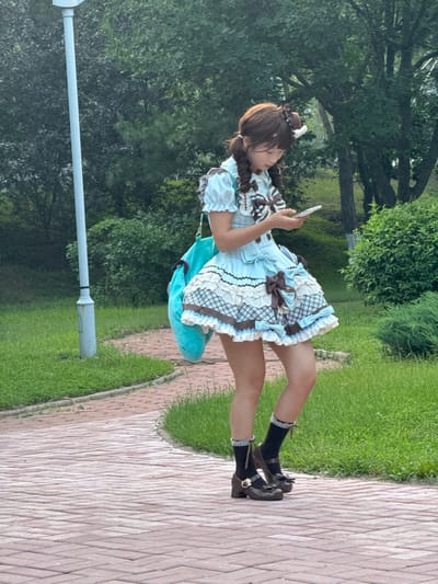 A young person in a blue and white Lolita-style dress with brown accents and bows is standing on a brick path in a park. They are looking down at a smartphone they are holding in their right hand. The person has brown hair styled in two braids and is wearing black socks with lace trim and brown, heeled shoes. A large, plush teal bag is slung over their left shoulder. The path is curved and surrounded by green grass and trees. A light pole is visible to the left of the person. The scene appears to be in Zhongtun, China.
