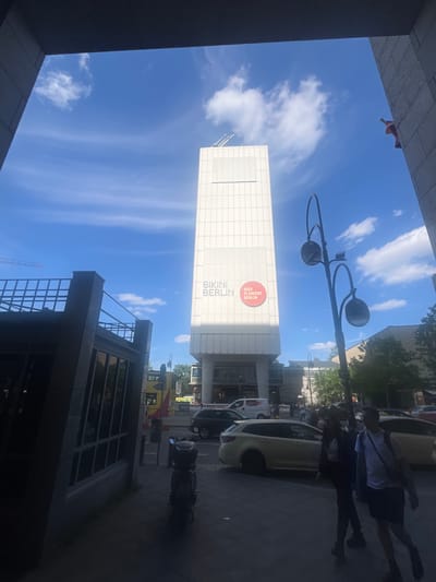 This is an outdoor, slightly upward-angled, full shot of a bright, sunny day in Berlin, Germany. The scene features a modern building with a white, grid-patterned facade that has "BIKINI BERLIN" written vertically on it. Above the text, a circular red logo with white text reads "HER FLANIERT BERLIN". A lamppost with two large black lights extends upwards into the blue sky dotted with wispy white clouds.
In the foreground on the right, a dark shadow is cast by a group of people, whose silhouettes are visible walking on the sidewalk. Their shadows are long, suggesting it's either morning or late afternoon. Next to them, a light-colored car is parked, and a dark motorcycle is also visible.
To the left and right of the frame, parts of buildings create a canyon-like effect, framing the sky and the central building. The overall impression is urban, with a focus on architecture and the interplay of light and shadow. The sky is clear and bright blue, indicating good weather conditions.
