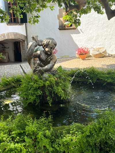 This image shows a stone fountain in Bunyola, Spain. A statue of a nude boy is positioned next to the fountain, with water jetting from it into the basin below. The fountain and surrounding area are covered in lush green vegetation. The building behind is white with architectural details such as arched windows. A flower pot with pink flowers hangs on the wall above the fountain. The water reflects the surrounding foliage.