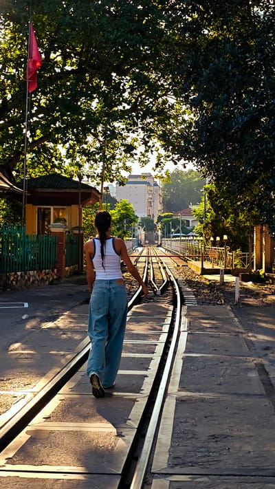 A woman walks down a railway track in Lao Cai Ward, Vietnam. She has dark hair braided and is wearing a white tank top and light blue jeans. The railway track is paved with concrete slabs between the rails. A small guardhouse with a red flag stands to the left of the tracks. Lush green trees and buildings line the sides of the railway. The lighting suggests it is either early morning or late afternoon.