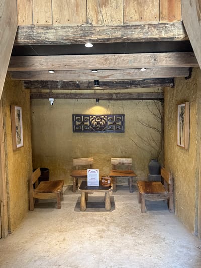 The image shows the interior of a building in Lũng Cú Commune, Vietnam. The walls are textured and yellowish-brown. There are wooden beams and planks along the left side, with embedded lights. A decorative metal panel hangs on the wall. Several small, rustic wooden stools and chairs are arranged in the room. Framed artworks are positioned high and low on the walls. A notice board stands on one of the stools.