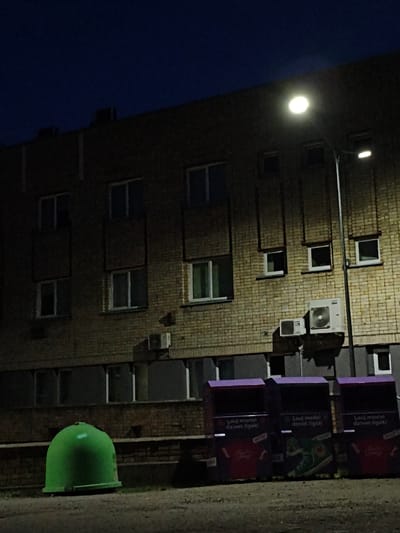 The image depicts a night scene in the city of Tukums, Latvia, focusing on the facade of a brick building. A street lamp illuminates the scene, casting a bright light on the building's exterior and a portion of the dark sky. The building features multiple windows, some of which are lit from within, while others are dark. Various air conditioning units are mounted on the brick wall. In the foreground, a bright green cylindrical trash receptacle is visible on the left, and three purple recycling bins are lined up on the right. These recycling bins are adorned with graphics and text. The ground in front of the building appears to be asphalt or concrete, with some debris and shadows present. The overall atmosphere is nocturnal and slightly urban.