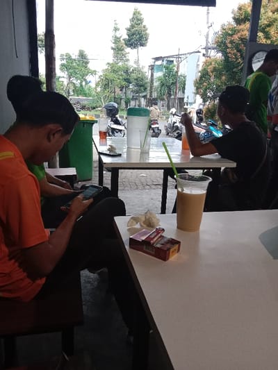 The image captures an outdoor or semi-outdoor cafe scene in Semarang, Indonesia. In the foreground, a person wearing an orange short-sleeved shirt and dark pants is seated on a dark wooden bench, actively using a black smartphone held in both hands. Next to this individual, a person in a green shirt is partially visible. On the white table immediately in front, a clear plastic cup containing a light brown beverage with a green straw is placed beside an open pack of "WIN" brand cigarettes, colored red and gold, and a crumpled white tissue.

In the middle ground, another person wearing a black t-shirt and a dark baseball cap is seated at a white table, largely facing away from the camera, drinking from a glass containing an orange liquid. On this table are additional items including a glass of dark amber liquid with a green straw, a large white water dispenser with a green lid, and another black smartphone. A green trash can with a yellow lid is positioned near this table.

The background reveals a paved ground, likely a street or sidewalk, bordered by various trees and foliage. Multiple motorcycles are parked and present on the street, including a black scooter with a helmet and a blue scooter. Several light-colored urban buildings are visible in the distance under an overcast sky. Other indistinct individuals are dispersed throughout the background.
