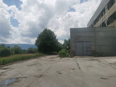 This daytime photograph, taken in Zhivkovo, Bulgaria, presents a blend of industrial decay and natural landscape under a cloudy sky. The foreground is dominated by a cracked and weathered concrete and asphalt surface, likely an old road or yard, with patches of dirt, small puddles, and visible tire tracks curving off to the left.
On the right, a large, austere concrete building stands prominently. It features a heavy, double-leaf metal door in a dull grey, suggesting a warehouse or industrial entrance. Above and behind this structure, the upper levels of a larger, multi-story industrial complex are visible. This building appears abandoned or in disrepair, characterized by numerous broken or missing windows and weathered concrete facades.
To the left of the scene, lush green vegetation, including tall grass, bushes, and trees, flourishes, creating a contrast with the concrete structures. In the far distance, a range of mountains can be seen, partially obscured by the greenery. The sky above is filled with large, bright white and grey clouds, with patches of blue sky visible, and a thin power line stretches horizontally across the frame. No explicit activity is taking place, and no discernible text is visible on any of the structures. The overall atmosphere conveys a sense of stillness and a landscape where nature slowly reclaims an aging industrial site.