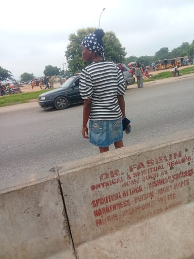 In Kagini, Nigeria, a young woman stands on a roadside barrier, her back to the viewer, wearing a striped t-shirt and a denim skirt. She holds a dark object in her right hand. The barrier is made of concrete and has red lettering that appears to advertise spiritual healing and business success, listing "SPIRITUAL ATTACK - BUSINESS SUCCESS" and "NEMENESS - POISON - WICCY MAPES" among other services. Behind her, a dark sedan is parked on the road, and various other vehicles, including multiple yellow auto-rickshaws, are visible in the background, along with trees and a cloudy sky. Several people are seen standing or walking around the vehicles, suggesting a public area or marketplace.
