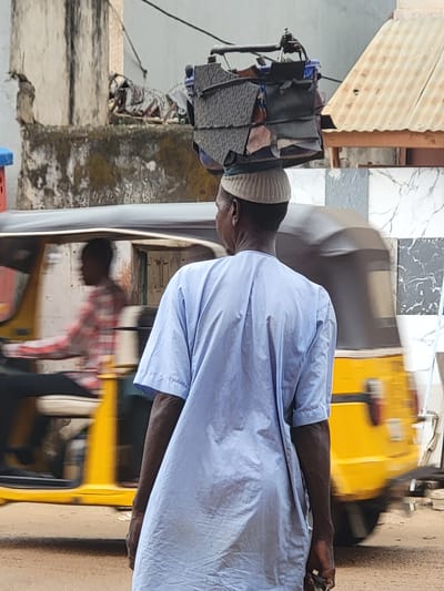This vertical image, set in Maiburiji, Nigeria, captures an outdoor street scene under bright daylight.
The main subject is a dark-skinned man, positioned in the center, facing left in profile from the waist up. He wears a light blue, long-sleeved garment, likely a `kaftan` or `bubu`, and a light-colored skullcap (`kufi`). His right arm is visible, extended forward, and his hand is holding a small, dark object, possibly a phone or remote, with a ring on one finger.
Behind the man, to his right (from the viewer's perspective), is a yellow three-wheeled vehicle, characteristic of an auto-rickshaw or `keke napep`. The vehicle appears blurred, suggesting either motion or a shallow depth of field. A reflection of another person, wearing a red and white plaid shirt, is visible in the vehicle's window or surface.
The background features various architectural and environmental details. To the man's left, a pile of assorted items, possibly bags or textiles, rests on a structure that might be part of a stall or another vehicle. Above this, a corrugated metal roof, light brown in color, is visible. The wall behind the yellow vehicle is covered with white and grey checkered tiles, some exhibiting marble-like veining. A sturdy brown pole or beam is also present in the background. The ground appears to be a rough, unpaved surface.
The overall scene conveys a typical daily activity or interaction in an urban or semi-urban setting. The bright, even lighting indicates it is daytime. There is no visible text in the image.