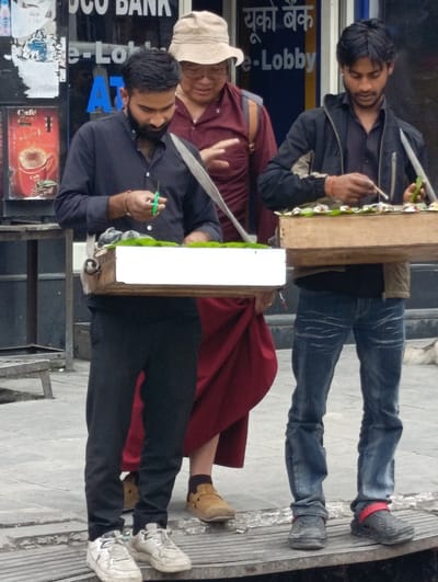 The image depicts a street scene in Manali, India, likely in a commercial area given the presence of a bank sign. Three individuals are visible. In the foreground, on the left, a man with a beard wearing a dark shirt and black pants is preparing what appears to be a food or tobacco product. He is holding a pair of green scissors in his right hand and a leaf in his left, poised over a wooden tray filled with ingredients. To his right stands another man in a black jacket and blue jeans, also attending to a similar tray. Behind them, a person in a light-colored hat and a maroon robe is partially visible. The environment appears to be outdoors, with a paved ground surface and a shopfront in the background displaying advertisements, including one for "Cafe". The overall impression is that of a marketplace or busy street where vendors are offering goods.
