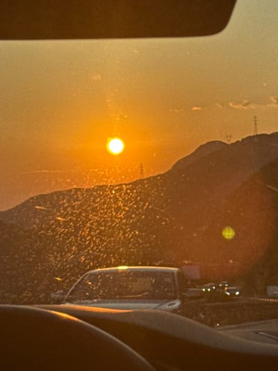 The image captures a sunset viewed from inside a vehicle. The sun, a bright, luminous orb, is positioned in the upper left quadrant of the sky, casting a warm orange glow across the scene. The sky is a gradient of orange and yellow, with a darker band across the top of the frame, possibly indicating the edge of the vehicle's interior.

In the midground, silhouetted mountains rise against the sunset. The slopes of the mountains are textured with numerous small, bright specks, likely caused by dust or insects on the windshield. A few faint clouds are visible near the horizon.

In the foreground, the dashboard of the vehicle is visible, along with part of the windshield. The windshield is covered in the same bright specks as the mountain slopes, creating a somewhat obscured view of the landscape.

Further in the background, on a road, several vehicles are visible. Their headlights are illuminated, suggesting it may be dusk or twilight. Power lines and poles are also visible on the ridge of the mountains. The overall atmosphere is serene, with the warm hues of the sunset creating a peaceful ambiance. The scene appears to be a common view during travel at sunset, particularly in a mountainous or rural area.