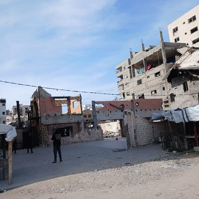 This image captures a scene of extensive urban destruction in Jabalia, Palestinian Territory, under a partly cloudy blue sky. The foreground consists of a dirt and gravel road leading into a wider, flat area of compacted earth scattered with debris. Three individuals are visible: a person in a black hoodie stands centrally, facing away from the camera, observing the damaged environment. Two additional figures are partially obscured in the left background, near a severely damaged building.
The midground is dominated by the skeletal remains of multiple structures. A prominent reddish-orange colored concrete building exhibits large missing sections of its facade, exposing interior spaces and structural elements, and bears numerous circular impact holes across its remaining walls. To its right, another building features a partially collapsed roof and extensive wall damage. A partial stone-clad wall with an arched opening stands on the right, also showing damage.
In the background, a multi-story concrete building, appearing either under construction or heavily damaged, shows exposed rebar, missing wall sections, and articles of laundry hanging from balconies or exposed floors. Other multi-story buildings are partially visible further in the background. A black utility line with a single light fixture stretches horizontally across the middle ground. On the far right, a makeshift market stall structure covered by white and blue tarpaulins is present.