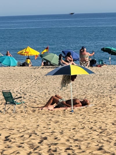 This media file depicts a sunny day at a beach in Quarteira, Portugal. The scene is populated by several people enjoying the beach, some relaxing under umbrellas and others sunbathing.
The subjects include multiple individuals, primarily adults, dressed in swimwear. There are several beach umbrellas of various colors, including green, blue, yellow, and turquoise, providing shade on the sandy shore. Beach chairs are also visible, one in a folded state and another in use. The setting is clearly a beach with fine sand and the blue ocean stretching into the distance. The bright sunlight suggests it is likely midday. The overall mood appears relaxed and leisurely, consistent with a typical beach day.
A notable detail is the yellow umbrella, which has text printed on it. The visible text reads "NADADOR SALVADOR", which translates to "Lifeguard" in Portuguese, indicating a safety service is present. The presence of numerous umbrellas and people scattered across the sand suggests a popular time at the beach. The calm water implies pleasant weather conditions.
