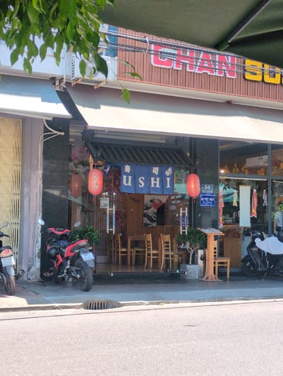 The image shows a vertical, eye-level, outdoor view of the front of a Japanese sushi restaurant in Quy Nhon Ward, Vietnam. The restaurant has a large, vertical, multi-colored sign, partially obscured by a green tree in the lower left. The sign reads “SUNG” at the top, followed by a series of Japanese characters, and “USHI” in Roman letters. The restaurant's entrance is decorated with Japanese lanterns and banners. Inside, wooden tables and chairs are visible. Two motorcycles are parked in front of the restaurant on the sidewalk. A sign that reads, “34” at the top with Vietnamese text below is affixed to the glass window. A concrete sidewalk and road are visible to the right.