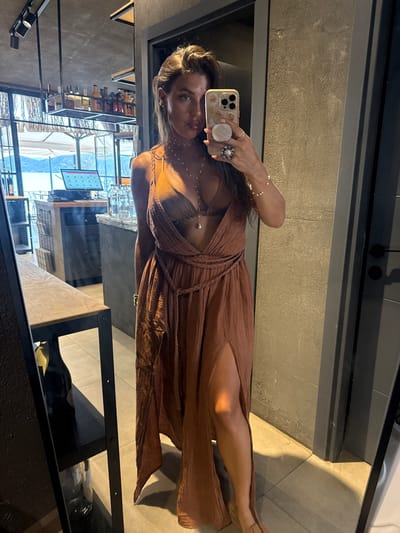 A woman stands in front of a mirror, taking a selfie. She is wearing a long, brown, flowy dress with a deep V-neck and a slit up the side, revealing her leg. She has on a bikini top underneath the dress, and several delicate necklaces. She is also wearing rings and a bracelet. Her hair is dark and pulled back. The background appears to be an indoor establishment, possibly a restaurant or bar, with shelves stocked with bottles. Through a large window in the background, a body of water and hills can be seen, suggesting a coastal location. The overall impression is that the photo was taken in Fethiye, Turkey, likely during a vacation.
