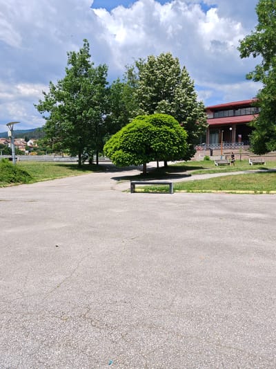 The image depicts an outdoor scene in Panagyurishte, Bulgaria, likely a public park or recreational area. A paved pathway occupies the foreground, with a grey bench placed in the middle ground. Beyond the pathway, a lush green lawn is visible, bordered by a yellow stripe. Several trees, varying in size and foliage, are scattered across the scene. One tree, with a distinct rounded canopy, stands prominently near the center. In the background, a building with a red roof and large windows is situated, and to the left, residential buildings and hills are visible. A lamppost stands on the left side of the image. Two individuals are present: one person, possibly a woman wearing dark clothing, walks on a path near the building, and another person is partially visible on a bench to the right. The sky is partly cloudy, with cumulus clouds against a blue expanse. The overall impression is one of a tranquil, daytime setting.