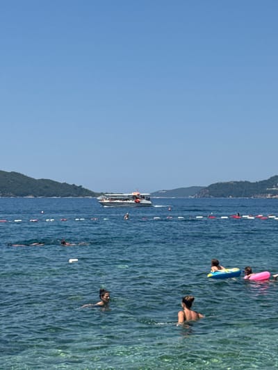 The media file depicts a sunny day at a beach in Kamenovo, Montenegro. The scene is split between a clear blue sky on the left and a vibrant blue sea on the right. The water is calm, with visible ripples and streaks of light reflecting off the surface.
Several people are enjoying the water. Some are swimming, while others are lounging on inflatable toys. In the foreground, a person is partially submerged, with only their head and shoulders visible above the water. Further out, a pink inflatable raft and a blue inflatable ring are being used by individuals. Closer to the shore, a boat with several people on deck is visible, suggesting a tour or a private vessel.
A line of buoys marks a boundary in the water, separating different areas of the sea. The shoreline is lined with lush green trees and vegetation, indicating a mountainous landscape behind the beach. The overall atmosphere appears relaxed and recreational, consistent with a typical beach day. The lighting suggests it is daytime, likely midday or early afternoon, with bright sunlight illuminating the scene. There is no visible text in the media file.