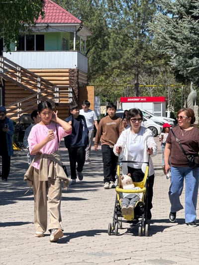 The image depicts a group of people walking on a paved pathway in Kenbulun, Kyrgyzstan. A woman pushes a child in a yellow stroller. A woman walks beside them. Several other individuals follow behind. The background features trees, a wooden structure, and a building with a red sign that says "BANKOMAT 24 часа". A white car is partially visible behind the stroller. One person is holding up two fingers.