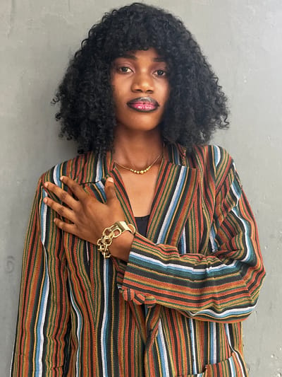 A Black woman with curly black hair is lying on a gray surface. She is wearing a striped blazer with orange, blue, white, and brown stripes, a gold necklace, and a large gold bracelet. She has dark lipstick and is looking towards the camera. The location is not specified.