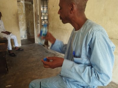 The image shows a man sitting on a floor in a room in Kaltungo, Nigeria, at coordinates 9.816, 11.315. He is wearing a blue long-sleeved shirt and is holding a plastic bottle with a blue cap in his right hand. He is looking down at the bottle and is about to open it with his left hand. Another man is sitting on the floor in the background wearing a white shirt. There is a wooden table on the left side of the image and a door in the background. The room is dimly lit and the floor is made of concrete.