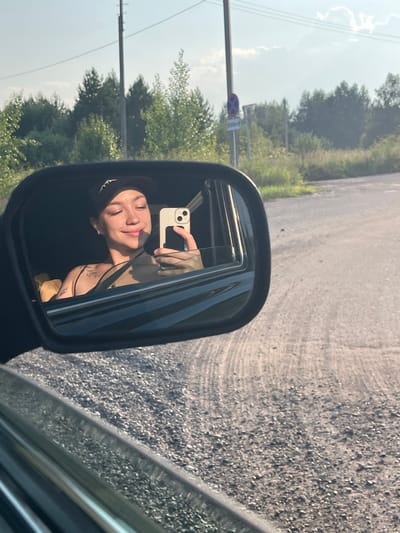 A young woman with light skin, dark hair, and wearing a black baseball cap is visible in the side mirror of a car. She is looking at her phone, which she is holding up to take a selfie. She has tattoos on her chest and arm. The car is on a gravel road, with trees and a cloudy sky in the background. There is a power line pole with wires on the left side of the frame, and a road sign with a red circle and a blue X is visible behind the trees on the right. The time of day appears to be late afternoon, as indicated by the warm light and long shadows. The location is not specified beyond being in Russia.