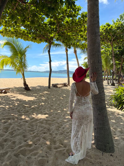 The image depicts a woman standing on a sandy beach in Ko Samui, Thailand, leaning against a palm tree. She is wearing a white lace dress with long sleeves and a red hat. The dress has an open back with pearl embellishments. Her left hand is resting on the palm tree. In the background, there is the ocean with a clear blue sky and some clouds. There are other palm trees and lush green vegetation surrounding the beach. The sand shows footprints and is partially shaded by the trees. Two logs are visible on the sand in the middle ground. The overall scene suggests a tropical beach setting.