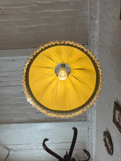 A low-angle shot depicts a mustard-colored lamp hanging from a textured white ceiling. The lamp shade is circular with gathered fabric and a decorative fringe. A lightbulb illuminates the space beneath the shade. The surrounding walls are painted white and appear to be constructed of brick. Two framed pictures are visible on the ceiling towards the top of the frame. A dark-colored decorative element appears on the right side of the frame. The location is Sarapul, Russia.