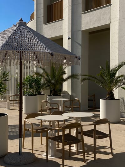 The image depicts an outdoor seating area, likely a cafe or restaurant, bathed in bright sunlight. In the foreground, a large, decorative umbrella with macrame detailing provides shade over several white, round tables and chairs with woven seats and wooden frames. The tables are simple and modern, with white cylindrical bases. Tall white planters containing palm trees and other lush greenery are strategically placed around the seating area.
In the background, a modern building with textured concrete walls and a railing on an upper level is visible. The architecture appears contemporary and clean. Shadows from the umbrella and furniture are cast across the tiled ground, indicating a sunny day. A security camera is mounted on the building's facade. The overall impression is one of a serene, upscale outdoor space. The context of "Sirius, Russia" implies this scene is located within that specific city. There are no people visible in the image, and no specific actions or interactions are taking place. The scene is static and captured from a slightly elevated perspective.