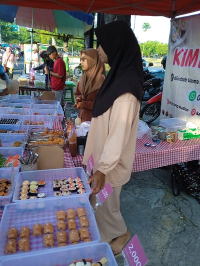 Here is a detailed description of the image:

**Scene:** A street food stall is set up on a sidewalk in what appears to be Madiun, Indonesia. The stall is under a red canopy. The stall sells Kimbab Umma, a Korean-style sushi.

**People:** Two women wearing hijabs stand behind the stall, presumably running the business. The woman closest to the camera wears a black hijab and beige clothing. A second woman behind her wears a brown hijab and coordinating clothing. A few other individuals are seen in the background, including a man wearing a red shirt and baseball cap, looking at a phone.

**Objects:** The stall has multiple clear plastic containers filled with various types of sushi. There are price tags next to some containers, indicating the cost of the sushi. A red and white checkered tablecloth covers the table. There are bottles of sauce, napkins, and other supplies on the table. A sign next to the stall promotes "Kimbab Umma," including contact information and a location "Depan OPRD Madiun." Motorcycles are parked behind the stall.

**Actions/Interactions:** The two women are standing behind the stall, possibly waiting for customers. Customers are browsing the sushi selection.

**Environment:** The scene is set outdoors on a sunny day. Trees and buildings can be seen in the background, indicating an urban area. The ground is paved.