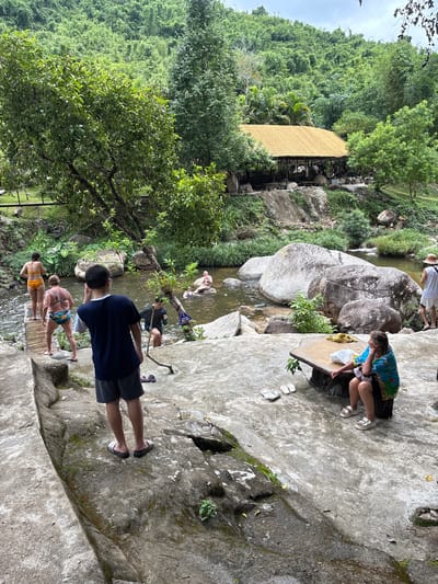This is a photograph of a resort near Khanh Vinh, Vietnam. The resort features a river with a sandy bottom and large rocks, surrounded by lush greenery and a bamboo-roofed building. Several people are visible in the image. In the foreground, a young person wearing a dark blue t-shirt and grey shorts stands facing away from the camera, looking towards the river. To the left of the river, a wooden walkway leads to a group of people. One woman in an orange bikini and another woman in a patterned swimsuit are walking on the walkway. Further into the river, several individuals are swimming or wading. On the right side of the frame, a woman in a blue and green sarong is seated at a rustic wooden table, with another person in a white shirt and shorts standing nearby. The environment appears to be a natural outdoor setting with a man-made resort infrastructure.