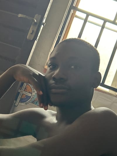 A young person with dark skin and a close-cropped haircut is positioned in the foreground, their face turned to the left, resting their chin on their left hand. The background features a closed door with a metallic handle and lock on the left, and a barred window with bright light filtering through on the right. A portion of a white T-shirt with a circular logo is visible on the person's chest. The lighting suggests an indoor setting, possibly in a residence, in Ihiagwa, Nigeria, given the overall ambiance and the presence of features common to Nigerian homes.
