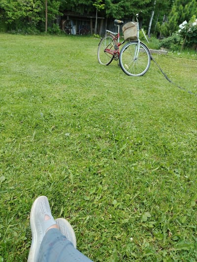 The image captures a serene outdoor scene in Novaya Prismara, Russia, depicting a grassy lawn with a bicycle parked in the background. In the foreground, a person's legs are visible, resting on the grass, clad in gray athletic shoes and blue pants. The bicycle, a red and silver model with a basket, is positioned on the lawn, its front wheel angled towards the right. The environment is characterized by lush green grass, scattered leaves, and trees in the background, suggesting a natural or semi-rural setting. The overall atmosphere is peaceful, with the sunlight illuminating the scene.