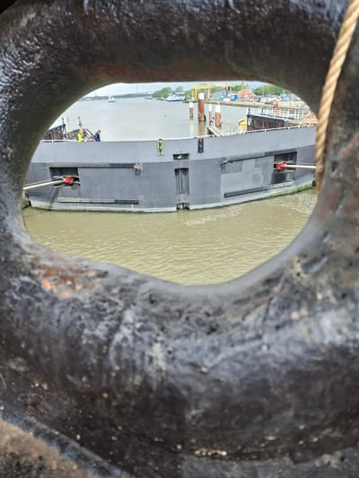 The image shows a view through a large, rusty metal ring. Beyond the ring, the view is of the Elbe River in Brunsbüttel, Germany. A cargo ship is docked along the riverbank. Several workers are visible on the ship. In the background, there are buildings and greenery characteristic of the town. The river is muddy-colored. A rope passes across the top left of the ring. The metal ring appears to be part of some larger structure or equipment.