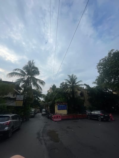 The image captures a street scene in Mumbai, India, under a cloudy sky. In the foreground, a wet asphalt road is visible, with a silver car parked on the left. Further down the road, a dark SUV is parked on the right, next to a yellow building with blue and white signage. Several trees, including palm trees, line the street, with lush greenery covering the sides of the buildings. There are also some traffic barriers and parked motorcycles visible. The sky is filled with dense, grey clouds, suggesting overcast or impending rain. The overall impression is a typical urban street in Mumbai, characterized by its vibrant vegetation and the presence of parked vehicles.